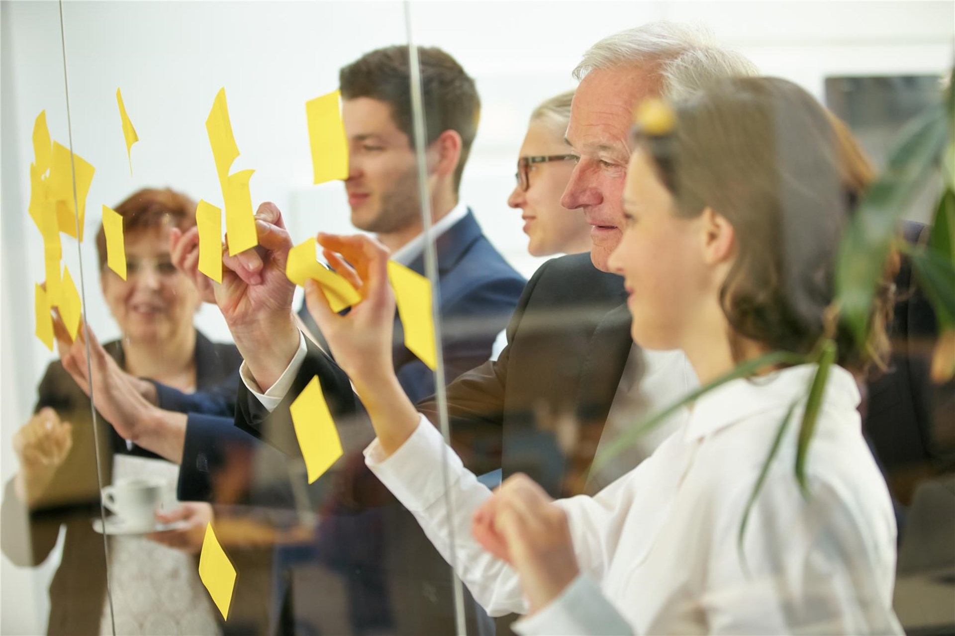 In einem Workshop stehen unterschiedlich alte Personen stehen hinter einer Glaswand und heften Post-Its an diese.